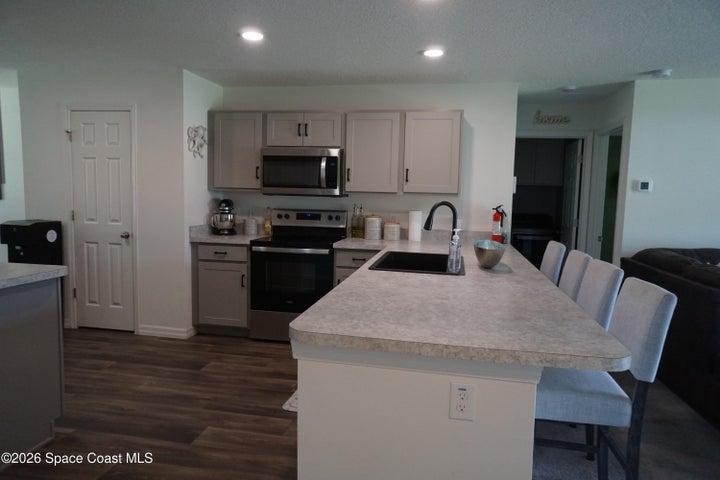 Open-concept kitchen featuring light-toned cabinetry with dark hardware, stainless steel appliances, a large center island with a gooseneck faucet, light-toned countertops, and vinyl plank flooring.