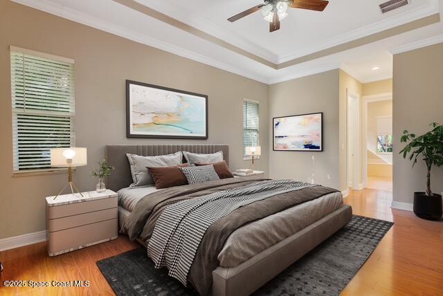 Bedroom with crown molding, light wood-style floors, a tray ceiling, ensuite bathroom, and a ceiling fan