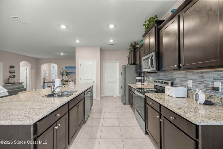 Kitchen featuring an expansive island with a double sink, granite countertops, dark wood cabinetry, a mosaic tile backsplash, and stainless steel appliances including a microwave, range, and refrigerator