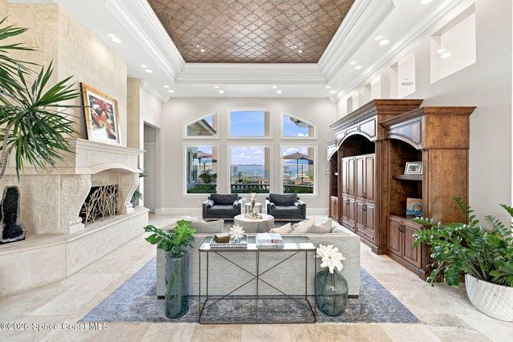 A showstopping great room with soaring ceilings, exquisite ceiling detail, a grand stone fireplace, custom cabinetry, and walls of windows showcasing spectacular Indian River views.