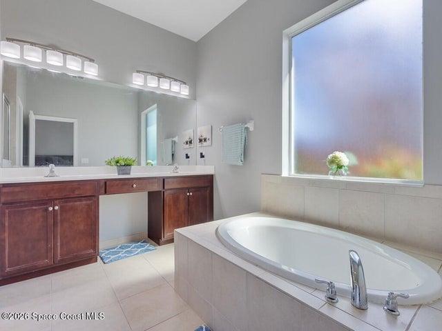 Soaking tub with tile surround, large privacy window, dual vanity with rich wood-finish cabinetry, extensive countertop space, and tiled flooring