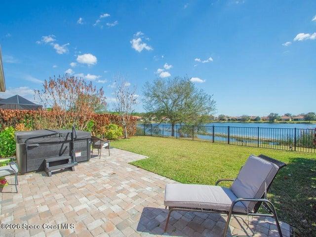 Spacious backyard featuring a paver patio with a built-in hot tub, lush green lawn, and a black wrought iron fence bordering a serene body of water