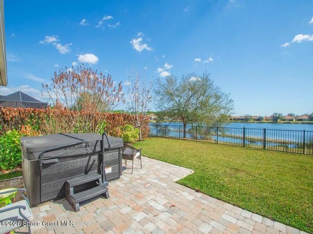 Spacious backyard featuring a paver patio with a built-in hot tub, lush green lawn, and a black wrought iron fence bordering a serene body of water
