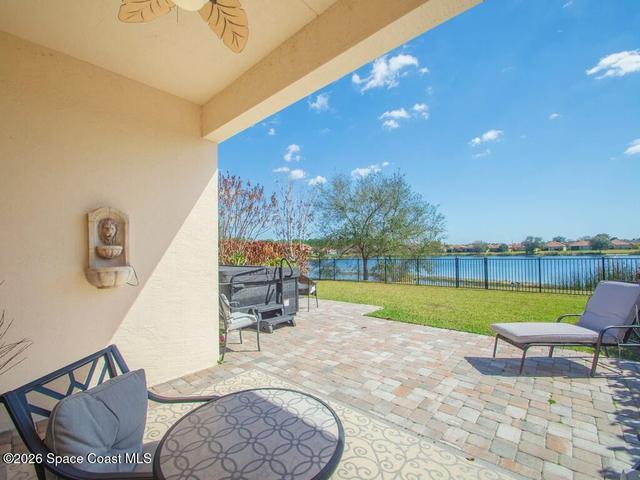 Covered patio with a ceiling fan, a wall-mounted water feature, and paver flooring overlooking a fenced lawn and a prominent lake