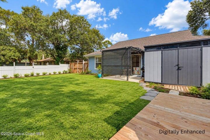 Expansive lawn with a white privacy fence, mature trees, wood decking, a covered patio, and an exterior storage shed